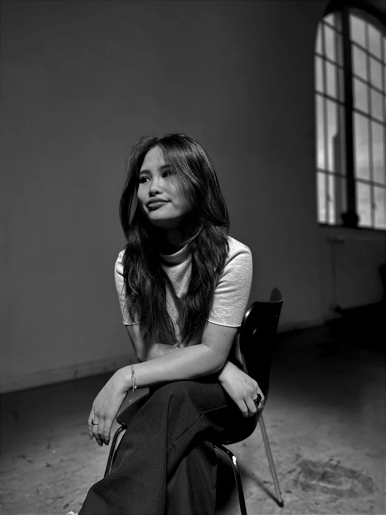 Romana Ta sitting on a chair in an empty room with tall windows, black and white photograph